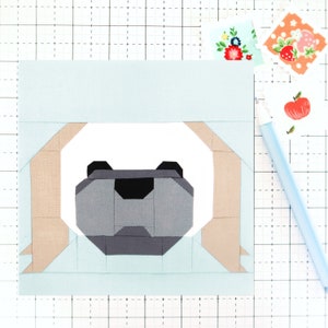 May include: A light blue and tan fabric block featuring a geometric bear face design. The bear's face is composed of gray, black, and white shapes. Decorative stickers and a light blue pen are in the background.