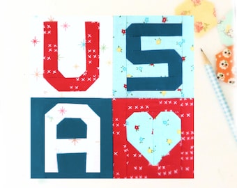 May include: A patchwork quilt block spelling out "USA" with a heart in the "A". The "U" is red with white stars, the "S" is blue on a floral background, the "A" is white on a blue background, and the heart is blue with a floral pattern on a red background.
