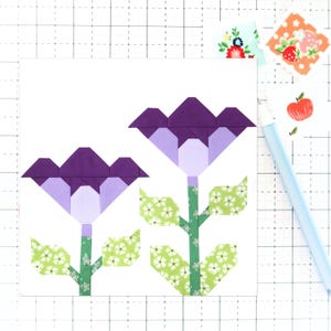 Puede incluir: Dos flores moradas y lavanda con hojas verdes sobre un fondo blanco. Las flores están hechas de tela y tienen un diseño geométrico.