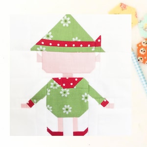 May include: A green and red patchwork elf applique with white flowers and red polka dots. The elf is wearing a green hat with a red band and white flowers.