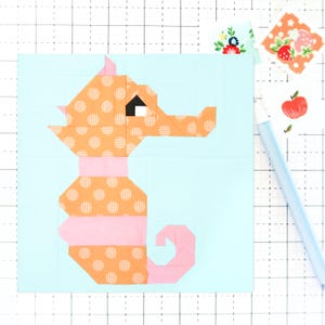 May include: A colorful seahorse quilt block. The seahorse is constructed from orange polka dot fabric with pink accents and a black eye. The background is light blue. Decorative paper and a light blue pen are in the background.