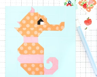Può includere: Un colorato blocco di quilt a forma di cavalluccio marino. Il cavalluccio marino è costruito con tessuto arancione a pois, con accenti rosa e un occhio nero. Lo sfondo è azzurro. Carta decorativa e una penna azzurra sullo sfondo.