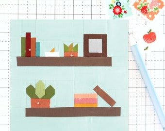May include: A light blue background with two brown shelves. The top shelf has a stack of books, a white candle, and a green plant in a brown pot. The bottom shelf has a green plant in a brown pot and a pink and orange box with a brown lid.