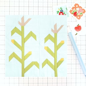 May include: A light blue fabric square with two green corn stalks with yellow kernels and pink tops. The fabric square is on a white grid background.