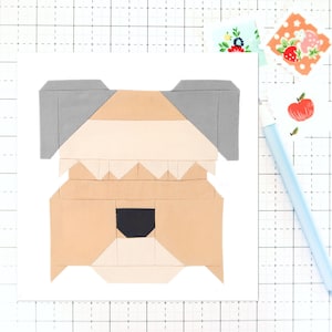 Puede incluir: Un bloque de colcha de retazos que representa la cara de un perro. El perro tiene pelo marrón, tostado y blanco, con orejas grises y una nariz negra. El bloque está hecho de varios cuadrados de tela cosidos juntos.