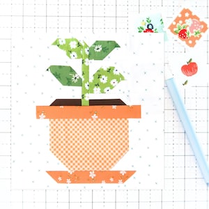 May include: A fabric art piece featuring a potted plant design. The plant has green leaves with white floral patterns, a brown pot, and an orange gingham pot. Additional fabric scraps and a light blue pen are also visible.