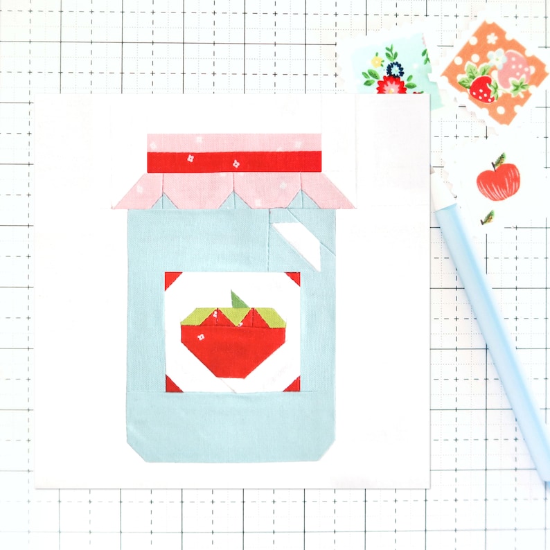 May include: A blue and white fabric patchwork quilt block depicting a jar of red apple jelly. The jar has a pink lid with white polka dots and a red band.