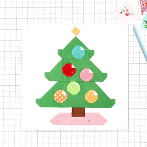 Op de afbeelding: Een geometrisch kerstboomontwerp in groen, versierd met kleurrijke, bedrukte ballen. De boom staat op een roze basis met een bruine stam. Een lichtblauwe potlood en bloemenstickers staan op de achtergrond, op een witte achtergrond.