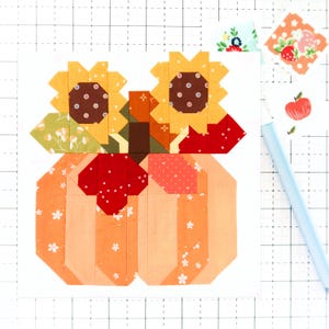 Op de afbeelding: Een patchwork quiltblok met een pompoen en twee zonnebloemen. De pompoen is oranje met witte bloemendetails. De zonnebloemen hebben gele bloemblaadjes en bruine centra. Stofresten en een blauwe pen staan op de achtergrond.