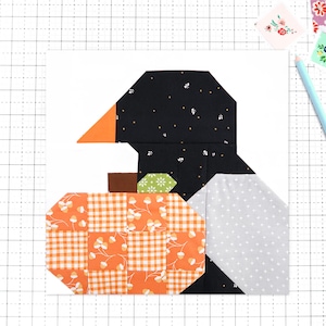 May include: A fabric quilt block pattern featuring a black bird with a bright orange beak, positioned next to a pumpkin made of orange and white checkered fabric with acorn prints, and a light gray wing with white polka dots.