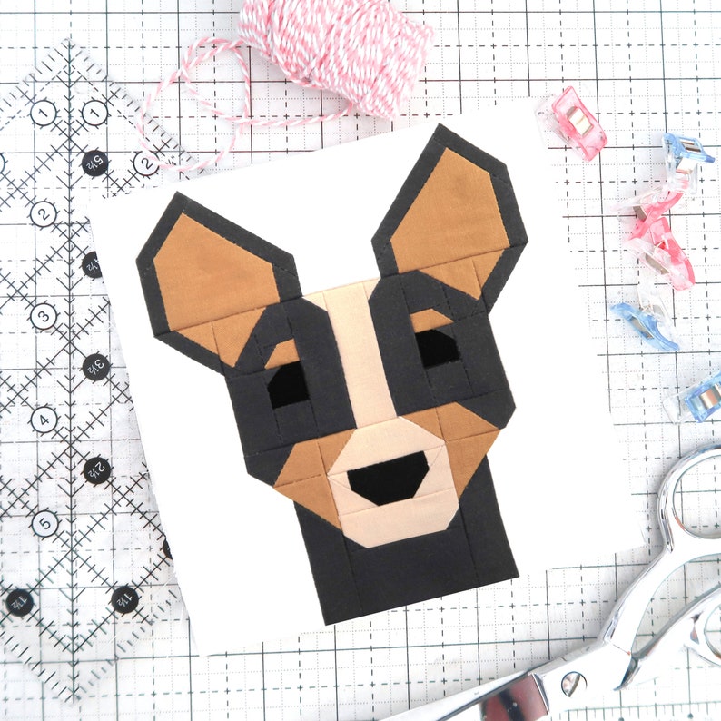 May include: A paper pieced pattern of a dog with brown, black, and white fabric. The pattern is on a white background and is surrounded by a grid.