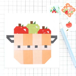 May include: A patchwork quilt block featuring a basket filled with red and green apples. The basket is made of orange and white fabric with black accents.