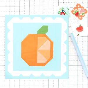 May include: A light blue square with a white scalloped border. Inside the square is a bright orange, segmented orange with a green stem and leaves. The orange is on a light blue background.