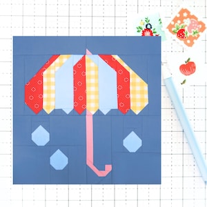 May include: A blue fabric square with a red, yellow, and blue striped umbrella with a pink handle and three blue raindrops.