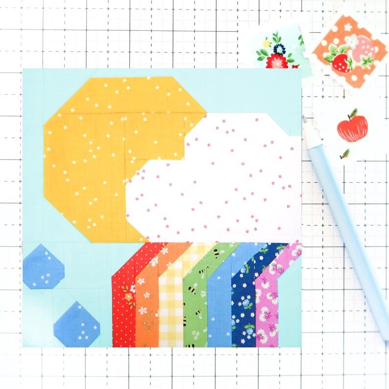 May include: A patchwork quilt block featuring a yellow sun, a white cloud with pink polka dots, and a rainbow made of colorful fabric strips. The background is light blue with two blue raindrops.