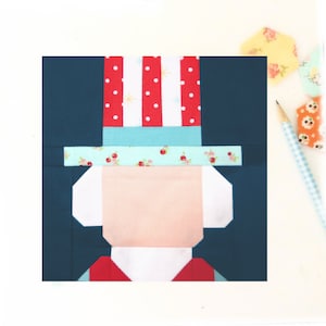 May include: A red, white, and blue fabric applique of a person wearing a hat. The hat has red and white stripes and white polka dots. The person's face is pink and the hat is on a dark blue background.
