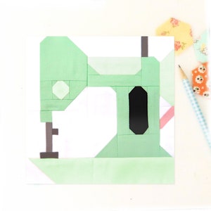 Könnte beinhalten: Ein grüner und weißer Patchwork-Quiltblock, der eine Nähmaschine darstellt. Die Nähmaschine hat eine schwarze Nadel und einen rosa Faden.