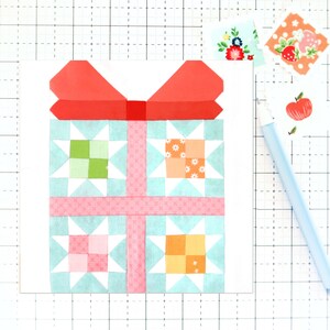 Puede incluir: Un bloque de colcha hecho a mano con un lazo rojo en la parte superior de una caja de regalo de patchwork azul y rosa. La caja de regalo está compuesta por cuatro cuadrados más pequeños, cada uno con un patrón diferente.