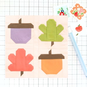 Może przedstawiać: Blok patchworkowej kołdry z jesiennym motywem. Blok składa się z czterech kwadratów, każdy z innym wzorem. Lewy górny kwadrat przedstawia fioletowy żołądź z brązową czapką. Prawy górny kwadrat przedstawia zielony liść. Lewy dolny kwadrat przedstawia pomarańczowy liść. Prawy dolny kwadrat przedstawia pomarańczowy żołądź z brązową czapką.