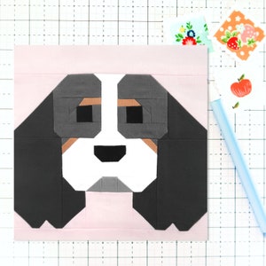 May include: A patchwork quilt block depicting a dog's face. The dog has black ears, a white muzzle, and grey eyes. The background is pink.