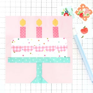 May include: A pink and white birthday cake with three yellow candles and a green base. The cake is decorated with pink and white checkered frosting and colorful sprinkles.