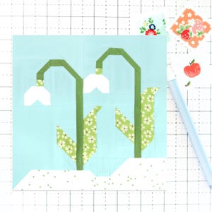 May include: A paper pieced quilt block featuring a design of two white snowdrops with green stems and leaves on a light blue background. The snowdrops are surrounded by a white background with small green dots.