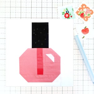 Op de afbeelding: Een roze en zwarte nagellak fles met een witte dop. De fles is gemaakt van stof en heeft een patchwork ontwerp.