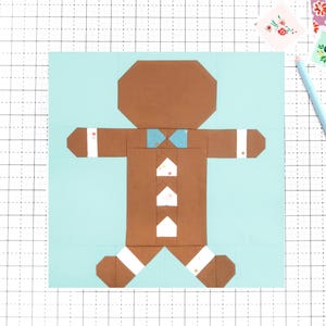 May include: A brown paper gingerbread man with a blue bow tie and white accents on a light blue background. The gingerbread man is made of folded paper and is on a grid surface. A light blue pencil and floral stickers are also present.
