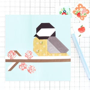 May include: A blue and white patchwork quilt block featuring a bird with a yellow, gray, and black body perched on a brown branch with pink flowers.