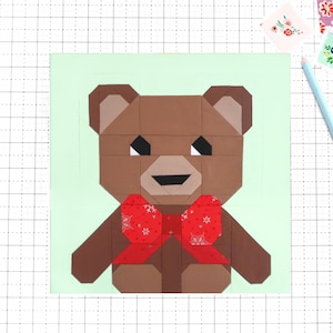 Op de afbeelding: Een geometrisch bruin teddybeerontwerp met een rode strik op een lichtgroene achtergrond. De beer heeft zwarte ogen en een lachende mond. Een lichtblauwe potlood en vierkante bloempatronen zijn ook zichtbaar.