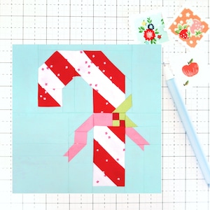 Op de afbeelding: Een rode en witte snoepstok met een roze strik en groene bladeren op een lichtblauwe achtergrond. De snoepstok is gemaakt van stof en is een patchwork-ontwerp.