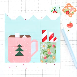 May include: A pink mug with a green Christmas tree design and a white rim, next to a green mug with a floral pattern and red and white striped straws. The mugs are on a light blue background with a white scalloped border.