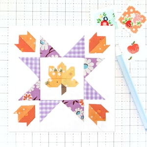 May include: A white fabric square with a purple and white gingham pattern, a floral pattern, and a yellow leaf design. The fabric square is surrounded by a white border.