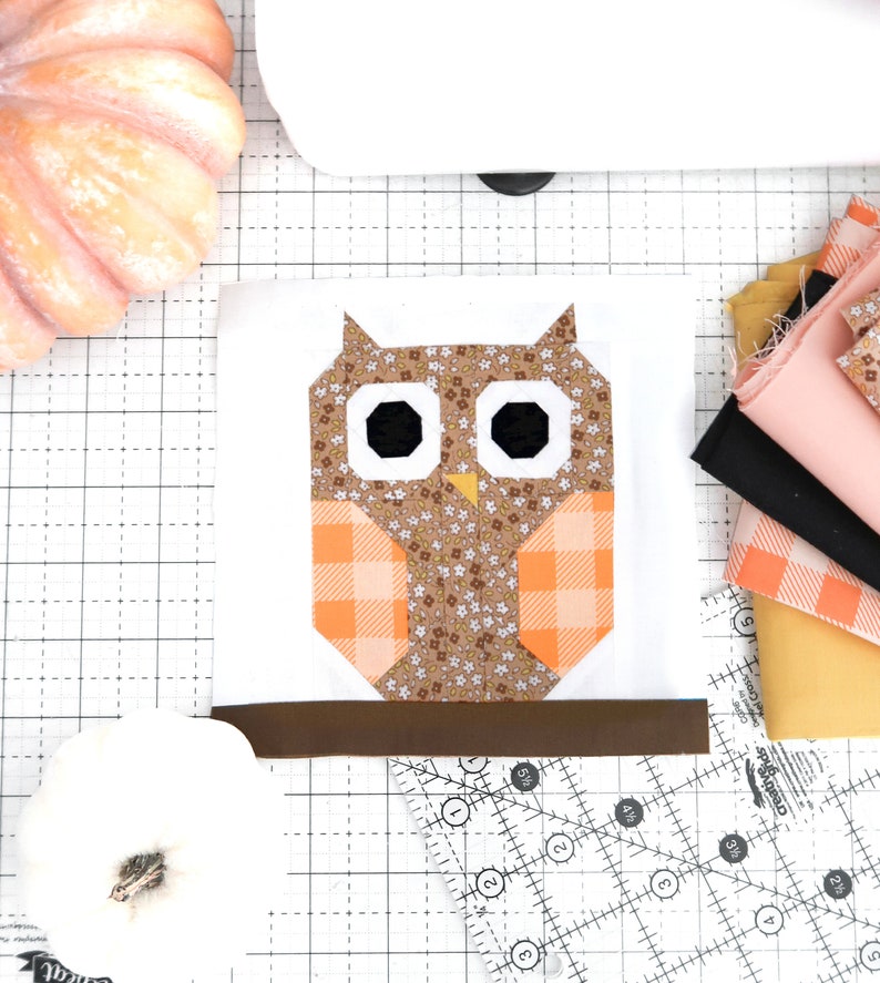 May include: A brown and orange patchwork owl applique on a white background. The owl has black eyes and a brown beak. The owl is sitting on a brown branch. The applique is on a white fabric with a grid pattern.