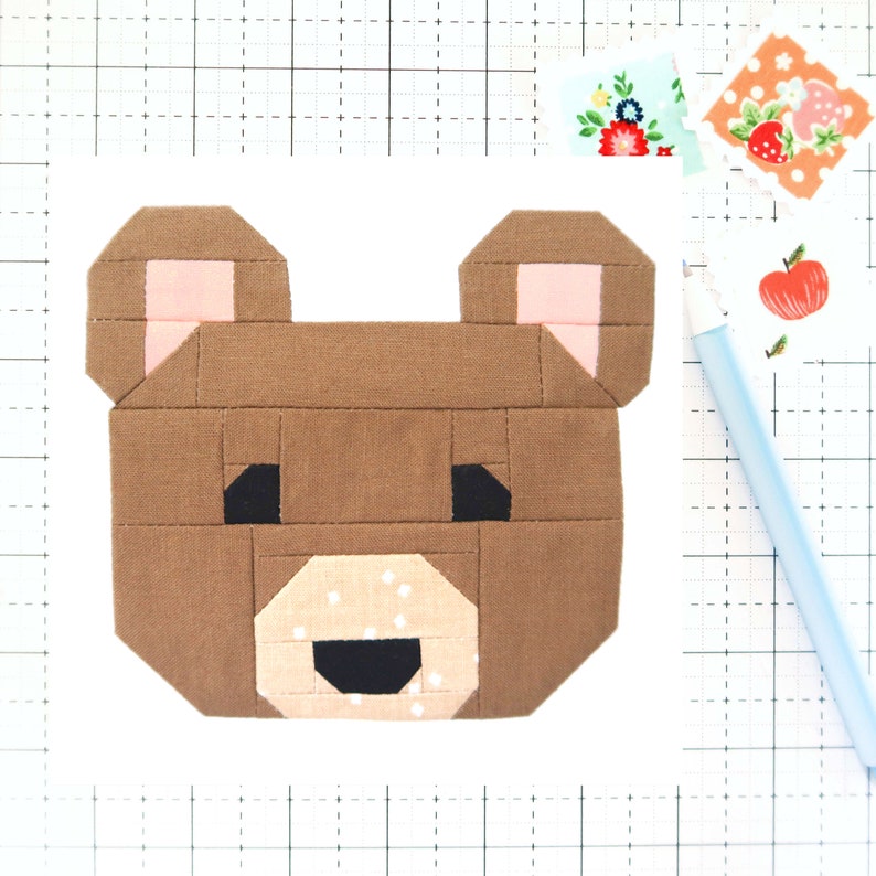 May include: A brown bear face made from fabric squares. The bear has pink ears, black eyes, and a black nose. The bear's mouth is open and has a white fabric with pink polka dots.
