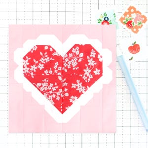 Puede incluir: Un cuadrado de tela rosa con un contorno blanco en forma de corazón. Dentro del corazón hay una tela roja con un patrón floral blanco.