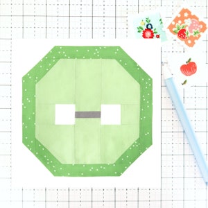 May include: A green octagon-shaped fabric button with white polka dots and a gray center. The button is on a white grid background.