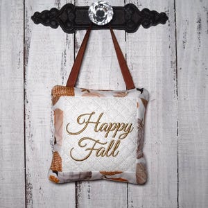 May include: A square fabric ornament featuring a white quilted center with the embroidered gold text Happy Fall. The border displays a pattern of pumpkins and autumn leaves in shades of orange and brown. It hangs from a brown ribbon.