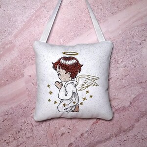 May include: A square white fabric ornament with a subtle glitter finish, featuring an embroidered design of a kneeling angel with brown hair, white wings, and a gold halo, surrounded by small gold stars, suspended by a white ribbon loop.