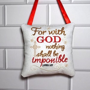 May include: A white square fabric ornament with a red ribbon hanger. The front features embroidered text reading For with GOD nothing shall be impossible LUKE 1:37 in red and gold thread, accompanied by a gold cross and small star accents.