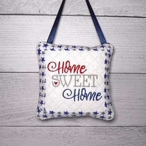 May include: A square quilted fabric ornament featuring the embroidered text Home SWEET Home with a small red heart. The white center is bordered by a pattern of blue and gray stars on a white background, finished with a dark blue ribbon hanger.