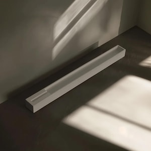 Puede incluir: Una bandeja larga y rectangular, de color blanco, con un canal empotrado. La bandeja está colocada sobre una superficie oscura, con sombras de una ventana u otra fuente de luz que crean un patrón en la escena. El diseño minimalista sugiere una estética moderna.