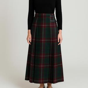 May include: A long, pleated skirt in a green and burgundy plaid pattern. The skirt has a high waist and is paired with a black long-sleeve top. The skirt features two decorative leather straps at the waist.