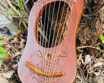 May include: A brown wooden lyre with a carved design of a figure playing a harp. The instrument has 10 strings and is set against a natural background of a tree trunk and foliage. The lyre is a musical instrument.