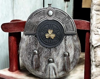 Könnte beinhalten: Ein traditioneller schottischer Sporran, eine Tasche, die zum Kilt getragen wird. Der Sporran hat einen silberfarbenen Metall-Cantle mit keltischen Knoten, eine schwarze Lederklappe mit einem goldfarbenen Kleeblatt und einen grauen Pelzkörper mit drei Quasten.
