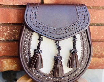 Op de afbeelding: Een bruine leren sporran met een wit middenpaneel en decoratieve kwastjes. De sporran heeft een klep met een geperforeerd ontwerp en is versierd met zilverkleurige studs rond de randen. Dit traditionele Schotse accessoire is ontworpen om met een kilt te worden gedragen.