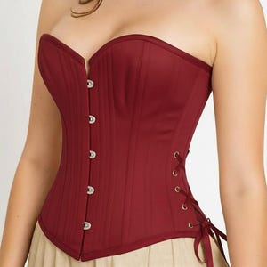 May include: A burgundy strapless corset with a sweetheart neckline. The bodice features vertical seams and silver-toned button closures down the front. The back has a lace-up design with burgundy ribbons.