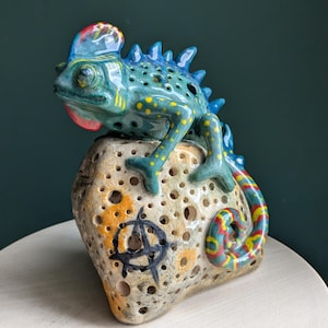 May include: A ceramic chameleon sculpture with a blue body, yellow spots, and a red crest, perched on a porous, beige, rock-like base. The base features a black circular symbol with an A inside, and the chameleon has a curled, multi-colored tail.