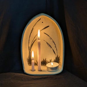 Puede incluir: Un portavelas de cerámica con borde azul claro, con tres velas encendidas. El soporte tiene un fondo beige con diseños de plantas pintadas en negro. También hay una pequeña vela de té encendida.