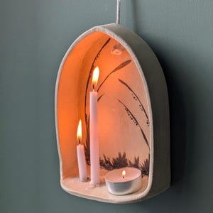 Könnte beinhalten: Ein bogenförmiger Wandkerzenhalter aus Keramik in hellem Beigeton. Das Innere ist mit einem handgemalten schwarzen botanischen Motiv verziert. Er enthält zwei dünne weiße Stabkerzen und ein rundes Teelicht, die alle brennend leuchten.
